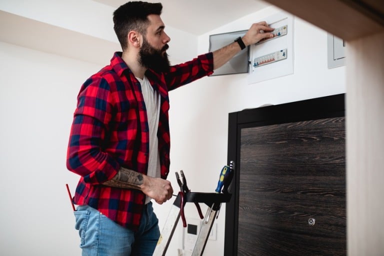 Best Electrical Panel Upgrade Electrician in West Palm Beach Florida Young bearded repairman standing on ladder and repairing fuse box.