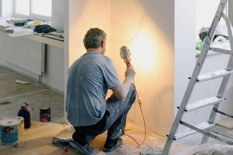 Best Home Remodel Electrician in West Palm Beach A Man Using a Light While Crouching Looking at a Corner in a Home with a Ladder in View and Wall Repair Tools on the Floor